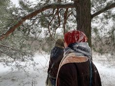 two people are standing in the snow near a tree and one is wearing a red head scarf