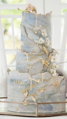 a three tiered blue and gold wedding cake with flowers on the top, in front of a window