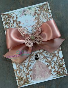 a pink and gold wedding card with a bow on it's front, sitting on a wooden surface