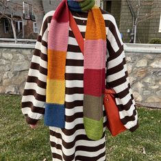 a woman standing in the grass wearing a multicolored sweater and scarf over her shoulders