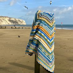 a crocheted blanket draped over a wooden post on the beach with seagulls flying in the background