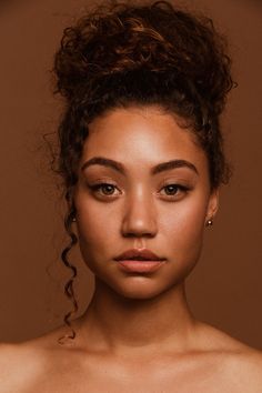 a young woman with curly hair and makeup looks at the camera as she stands in front of a brown background