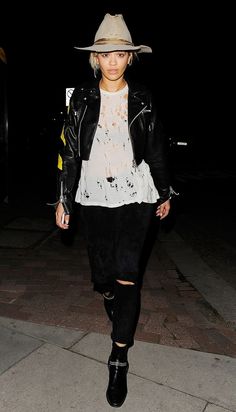 a woman wearing a hat and black pants is walking down the street at night with her hand in her pocket