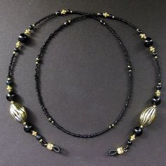 "Inky black beads make up the color of this eyeglass chain (or badge lanyard) when combined with the warm shine of gold plated accents including two large swirl textured egg shape beads. All together creating a beautiful and classy style for this handmade eyeglass chain, face mask holder, or badge lanyard. The length of the strand is made with smooth beads at the nape for the comfort of the wearer and decorative beads at the ends to show off a sense of style. 29.5\" from tip to tip - 75 cm Lengt Black Glass Beaded Necklaces With Beaded Chain, Black Glass Beaded Necklace With Beaded Chain, Black Glass Beaded Necklace, Adjustable Black Necklace With Gold Beads, Adjustable Beaded Black Glasses Chains, Adjustable Black Beaded Glasses Chains, Elegant Black Glasses Chains For Party, Adjustable Black Beaded Necklaces As Fashion Accessory, Adjustable Black Beaded Necklace As Fashion Accessory