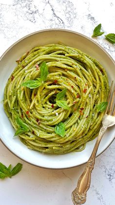 a white bowl filled with pesto pasta on top of a marble table next to silverware