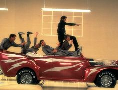 three men sitting on top of a red car in the middle of a showroom