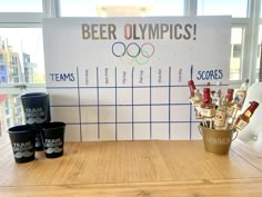 a wooden table topped with cups filled with wine and beer next to a sign that says beer olympics