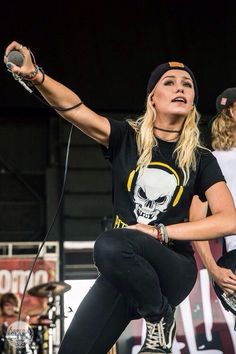 a woman on stage with her hands in the air while holding a microphone and wearing a skull t - shirt