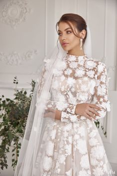 a woman in a white wedding dress with flowers on the skirt and veil, standing next to a plant