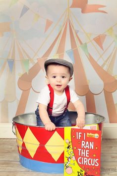 a baby sitting in a bucket with a book on the floor and a circus tent behind it