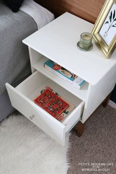 a white nightstand with books on it next to a bed and a framed picture in the corner