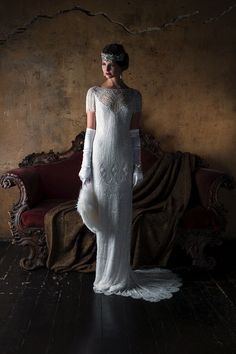 a woman in a long white dress with feathers on her head and gloves, standing next to a couch