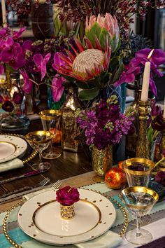 the table is set with plates, silverware and purple flowers in gold vases