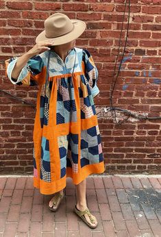 a woman standing in front of a brick wall wearing an orange and blue patchwork dress