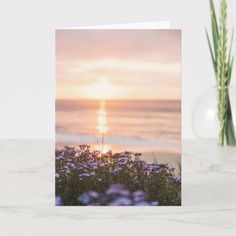 a greeting card with the sun setting over the ocean and flowers in front of it