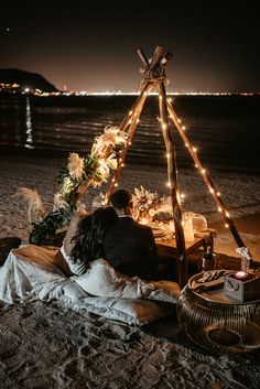 a man and woman are sitting in front of a teepee on the beach at night