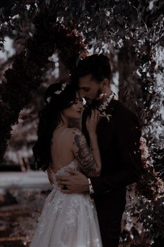 a bride and groom standing under a tree in their wedding day attire with tattoos on their arms