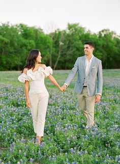 a man and woman holding hands walking through a field of bluebonnets with trees in the background