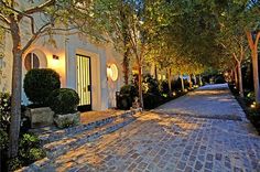 a cobblestone street lined with trees and bushes next to a white building at night