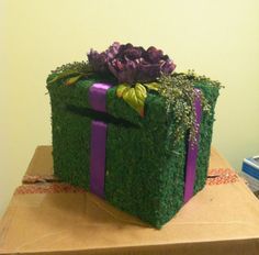 a green gift box covered in grass with purple ribbon and flowers sitting on top of it