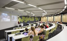 people are sitting at tables in a lecture hall with projector screens on the wall