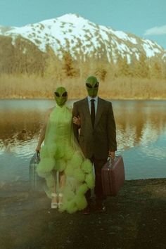 two people in alien costumes holding suitcases and standing next to a body of water