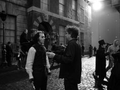 two people standing in front of a building talking to each other on a cobblestone street