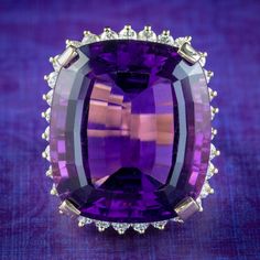 Boasting an enormous cushion cut amethyst, this spectacular cocktail ring was made to be seen! The amethyst weighs an impressive 40 carats (approx.) and has a luxurious, deep purple hue and glistening facets, complemented beautifully by a frame of sparkling brilliant cut diamonds around the border (approx. 1.55ct total).  Amethyst is the birthstone of February. It's considered a stone of protection, clarity and tranquillity and has been highly esteemed throughout the ages for its stunning beauty Luxury Cushion Cut Amethyst Ring For Formal Occasions, Luxury Amethyst Cushion Cut Ring For Formal Occasions, Luxury Amethyst Cushion Cut Ring For Formal Events, Cushion Cut Amethyst Ring For Formal Occasions, Cushion Cut Purple Amethyst Ring, Cushion Cut Amethyst Ring In Purple, British Crown Jewels, Colored Stone Rings, Edwardian Jewelry