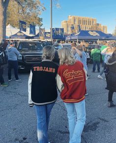 two people walking down the street in front of some parked cars and tents at an outdoor event