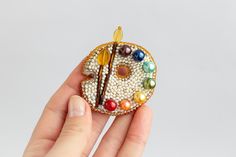 a hand holding a small beaded brooch with paintbrushes and beads on it