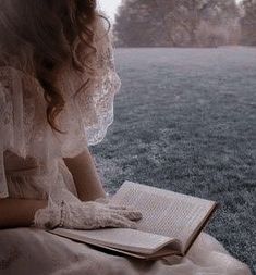 a woman is sitting on the ground reading a book and looking out over an open field