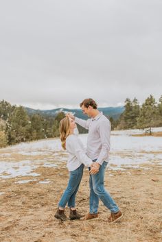 an engaged couple holding hands and walking through the snow in front of trees on a cloudy day