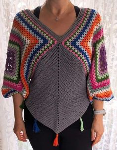 a woman standing in front of a window wearing a crocheted sweater with tassels