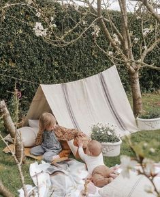 two children are sitting in the grass near a teepee and some trees with white flowers