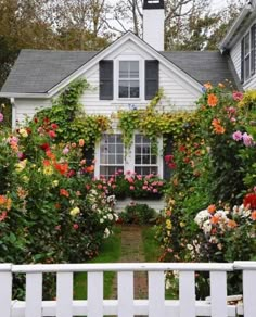 a white house surrounded by flowers and greenery with a white picket fence in front