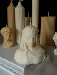 some candles that are sitting on a table with statues in the background and one candle has a woman's head on it