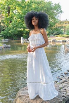 a woman standing on top of a rock next to a pond wearing a white dress