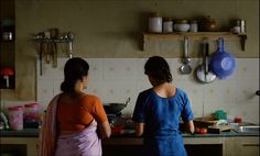 two women standing in a kitchen preparing food on the stove top, with pots and pans hanging above them