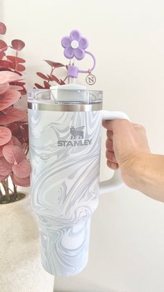 a person holding a white coffee cup with purple flowers in the background and a pink plant behind it