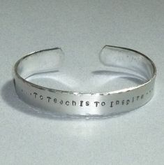 a silver cuff bracelet with the words to teach to inspire written on it, sitting on a white surface