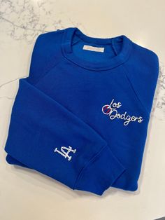 Whether you're a die-hard Dodgers fan or rooting for another team, this sweatshirt is the perfect addition to your game day wardrobe. Perfect for opening week festivities at the stadium or cozying up at home while cheering on your favorite team. This raglan crewneck sweatshirt is crafted from premium materials and embroidered with white, ecru and red thread.  Gear up for the season in style and comfort with a custom sweatshirt - the ultimate statement piece for any sports enthusiast. *Model is w Sporty Blue Sweatshirt With Embroidered Logo, Varsity Sweatshirt With Embroidered Logo For Sports Events, Blue Crew Sweatshirt For Game Day, Blue Sports Top With Logo, Blue Crew Neck Sweatshirt For Game Day, Team-colored Sweatshirt With Embroidered Logo For Sports Events, Collegiate Blue Top With Embroidered Logo, Blue Team Spirit Sweatshirt With Team Name, Blue Sports Sweatshirt With Embroidered Logo