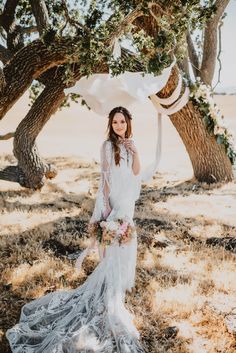 a woman standing in front of some trees wearing a white dress and holding a bouquet