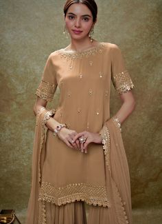 This exquisite Beige Georgette Kurta Sharara Set is a perfect blend of elegance and tradition. Made from georgette fabric, the beige kurta is adorned with delicate embroidery featuring mirrors, gota, sequins, and pearls, creating a stunning visual appeal. The neckline, hem, and sleeves are intricately embellished, complemented by small floral buttis that add a touch of grace. Teamed with a tiered sharara that showcases scattered mirror work, this ensemble exudes sophistication. The look is completed with a matching net dupatta, beautifully bordered and dotted with buttis, making it an ideal choice for Home Pooja, family gathering or festive occasions. Composition : Kurta and Sharara - Viscose Georgette and Dupatta - Soft net Care: Dry Clean Only and Vacuum Storage This product can be custo Mirror Work Kurta, Bridal Crop Top, Beige Kurta, Kaftan Kurta, Georgette Kurta, Kurta Lehenga, Prince Coat, Kurta Sharara Set, Kurta Sharara