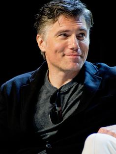 a man in black jacket sitting down with his hands on his hips and looking at the camera