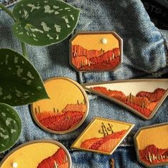 four embroidered patches with mountains on them sitting next to a green potted plant in the background