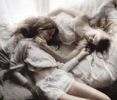 two women laying in bed with white sheets and pillows on top of each other, one woman has her arm around the other