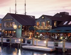 people are sitting at tables on the dock in front of some buildings with lights and umbrellas