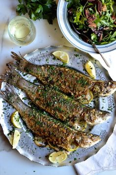 grilled fish with herbs and lemons on a plate next to a bowl of salad