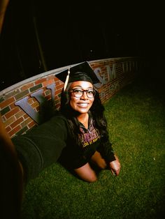 a woman wearing glasses and a graduation cap sitting on the grass in front of a brick wall