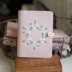 a pink notebook with embroidered flowers on it sitting on top of a wooden box next to a wire basket